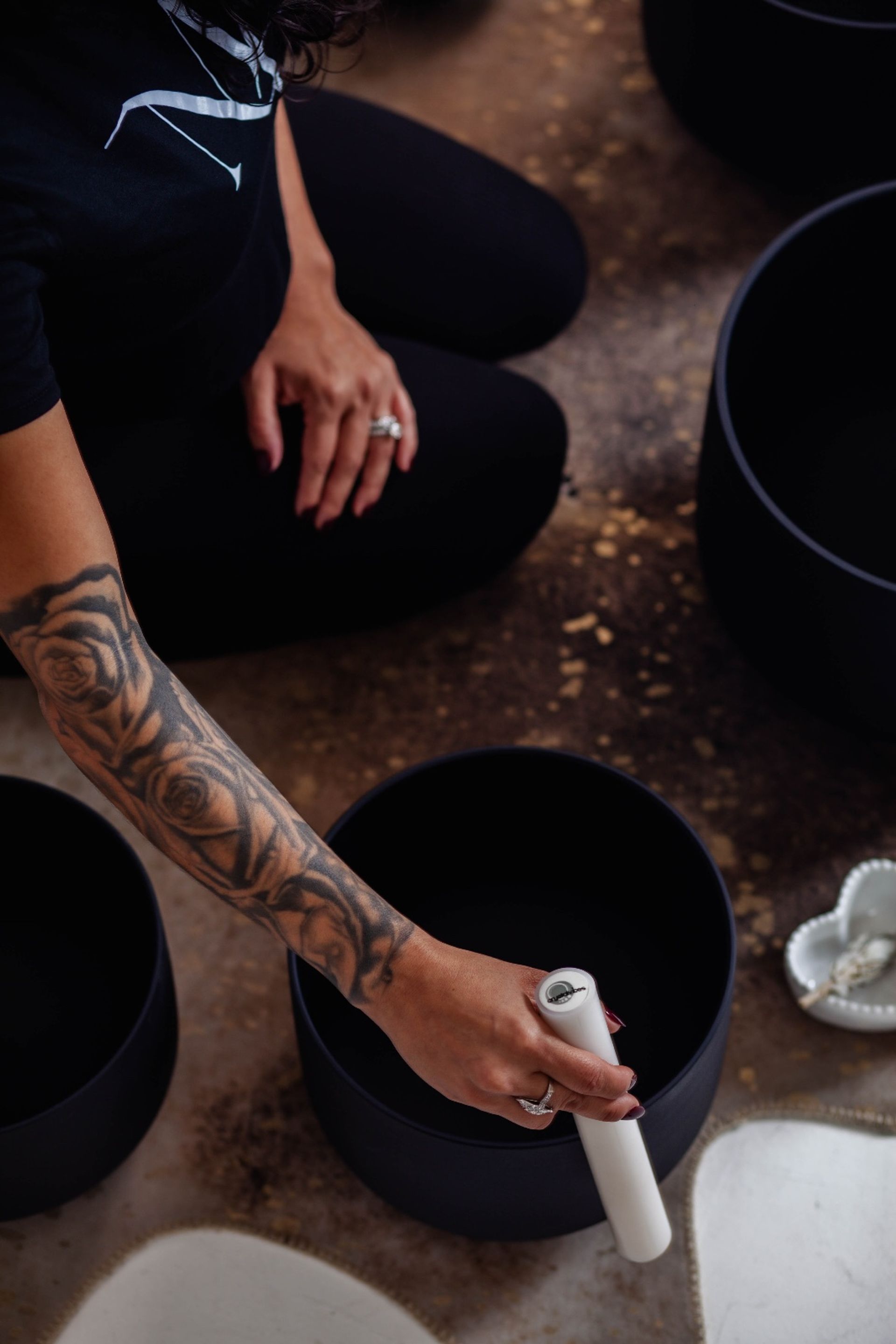 A tattooed arm kneeling on a studio floor strikes a black singing bowl with a white mallet during a sound-bath meditation.