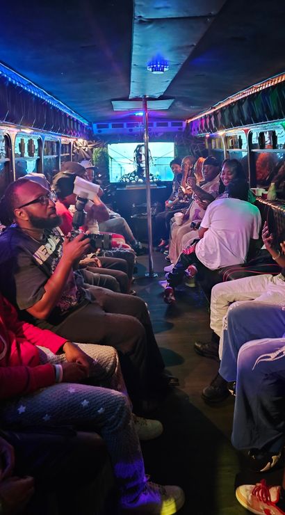 LED-lit party bus interior at night with people seated along both sides, colorful ambient lights and a center dance pole, a photographer holding a camera, a rear video screen, and passengers laughing and using phones.