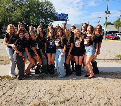Cheerful group photo of about 18 young women posing outdoors in coordinated black graphic T‑shirts, denim and boots on a sunny day in a sandy parking lot with trees and a bright blue sky.