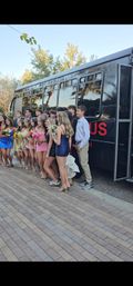 Group of teens in formal prom dresses and suits holding bouquets and corsages, posing outside a black party bus on a tree-lined suburban street with a brick-paved sidewalk.