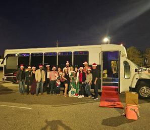 Large group of adults in Santa hats and festive outfits posing outside a white party bus with a red carpet in a parking lot at night — holiday party photo.