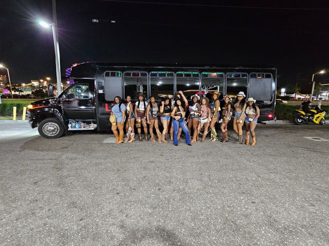 Nighttime group of women in cowgirl outfits posing in front of a long black party bus in a lit urban parking lot with city lights in the background