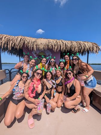 Group of women in swimsuits wearing green antenna headbands and colorful leis posing on a tiki-roofed party boat over calm blue coastal waters on a sunny day — tropical party boat cruise vibe