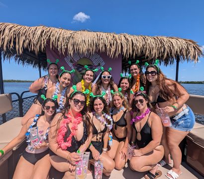 Group of women in swimsuits wearing green antenna headbands and colorful leis posing on a tiki-roofed party boat over calm blue coastal waters on a sunny day — tropical party boat cruise vibe