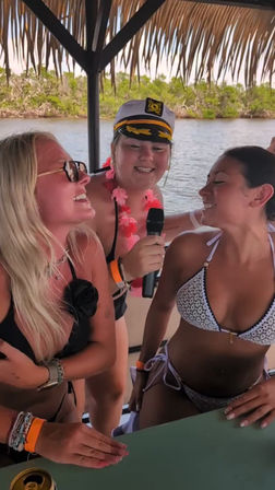 Three women at a tropical boat party under a thatched canopy — one wearing a captain's hat and pink lei sings into a microphone while two friends in bikinis laugh, water and mangroves in the background.