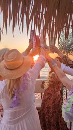 Friends toasting with colorful tumblers at a tropical sunset boat party, wearing leis, a straw sun hat and summer dresses beneath a thatched palapa as the sun sets over calm ocean waters.