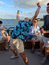 Friends partying on a boat at a sunny waterfront — man in a blue tropical shirt throws his arms up holding sunglasses while others laugh and sip canned drinks, sailboats and shoreline in the background.