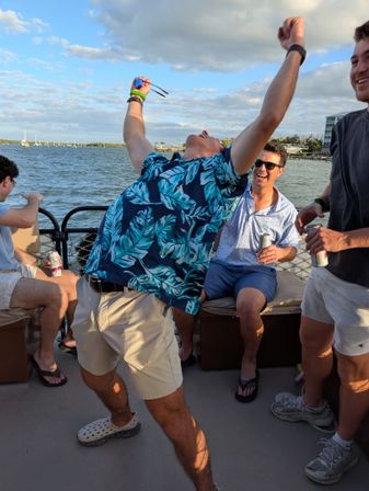 Friends partying on a boat at a sunny waterfront — man in a blue tropical shirt throws his arms up holding sunglasses while others laugh and sip canned drinks, sailboats and shoreline in the background.