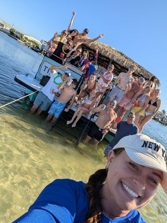 Selfie of a smiling woman with a lively tiki-style pontoon boat party behind her — friends in swimsuits and leis on the boat and knee‑deep in clear shallow coastal water on a sunny day.