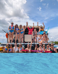 Tiki-themed pontoon party with a group in swimsuits posing on and around a decked boat in bright blue coastal water under a sunny sky