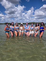 Eight friends in blue swimsuits standing in shallow coastal water on a sunny day, wearing heart-shaped sunglasses and leis and holding canned drinks under a bright blue sky with puffy clouds.