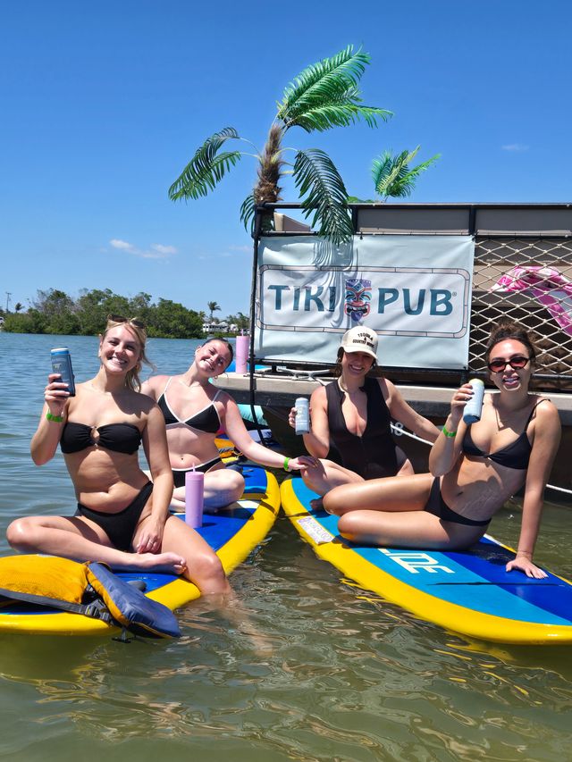 Four smiling women on paddleboards, toasting canned drinks beside a tiki‑themed floating bar with faux palm fronds on a sunny coastal inlet with mangroves