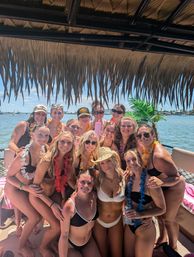 Group of smiling women in swimsuits wearing colorful leis on a tiki‑roofed party boat in a sunny coastal bay, palm accents and boats visible in the background.