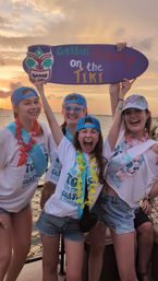 Four friends on a sunset waterfront cruise holding a purple sign reading Gettin' Freaky on the Tiki, wearing blue caps, leis, matching party shirts and denim shorts — tropical celebratory vibe.