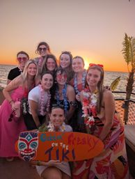Smiling group of women wearing leis on a tiki-themed boat at ocean sunset, holding a colorful surfboard sign that reads Gettin' Freaky on the Tiki