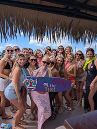 Group of women in swimsuits and leis under a thatched tiki roof on a sunny boat, smiling and holding a colorful “Gettin' Freaky on the Tiki” sign — tropical boat party scene