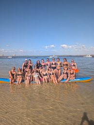 Smiling group of women in bikinis posing on a large paddleboard and in shallow coastal water, with boats nearby under a clear blue sky.