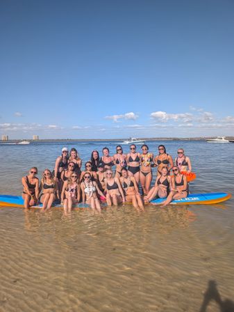 Smiling group of women in bikinis posing on a large paddleboard and in shallow coastal water, with boats nearby under a clear blue sky.