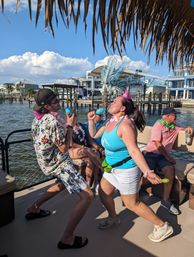 Two people dancing and shaking colorful maracas on a festive boat deck at a sunny waterfront marina, wearing leis and party hats under a thatched canopy with docks and coastal homes in the background.