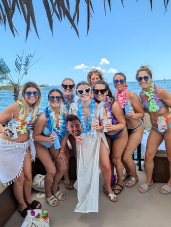 Group of women in swimsuits and sunglasses wearing colorful leis on a sunny tropical boat over blue ocean waters, holding canned drinks and a novelty cutout head beneath palm-frond shade