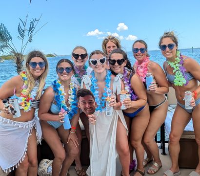 Group of women in swimsuits and sunglasses wearing colorful leis on a sunny tropical boat over blue ocean waters, holding canned drinks and a novelty cutout head beneath palm-frond shade