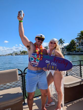Couple partying on a boat at a sunny waterfront — shirtless man in a captain's hat and lei raising a beer can, woman in a white swimsuit and lace cover-up holding a colorful tiki sign, palm trees, marina and blue sky in the background.