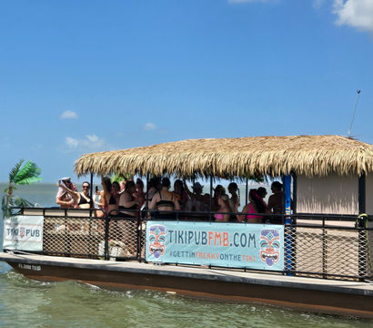 Tiki-style party boat with thatched roof and people in swimsuits cruising calm coastal Florida waters under a bright blue sky