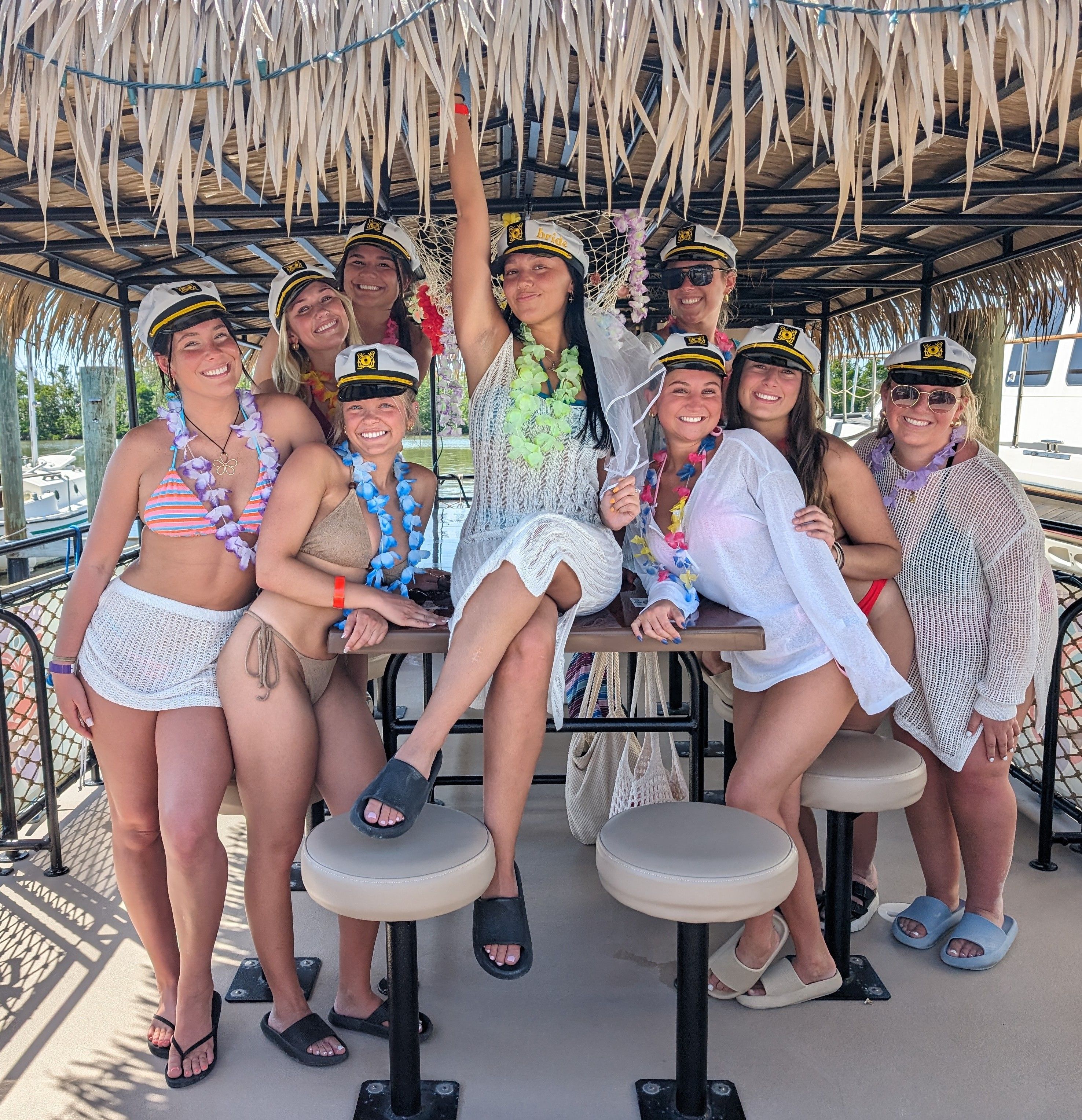 Bachelorette party group of smiling women in captain hats and colorful leis on a tiki-style party boat at a marina, wearing swimsuits and cover-ups under a thatched roof