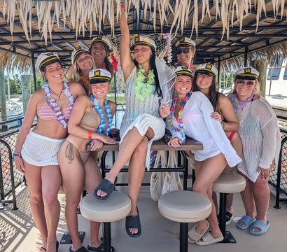 Bachelorette party group of smiling women in captain hats and colorful leis on a tiki-style party boat at a marina, wearing swimsuits and cover-ups under a thatched roof