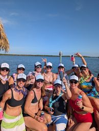 Smiling group of women in matching "Girls Trip" trucker hats, sunglasses and leis, wearing swimsuits and holding drinks while posing on a boat by a calm blue bay under a clear sunny sky.