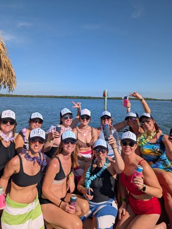 Smiling group of women in matching "Girls Trip" trucker hats, sunglasses and leis, wearing swimsuits and holding drinks while posing on a boat by a calm blue bay under a clear sunny sky.