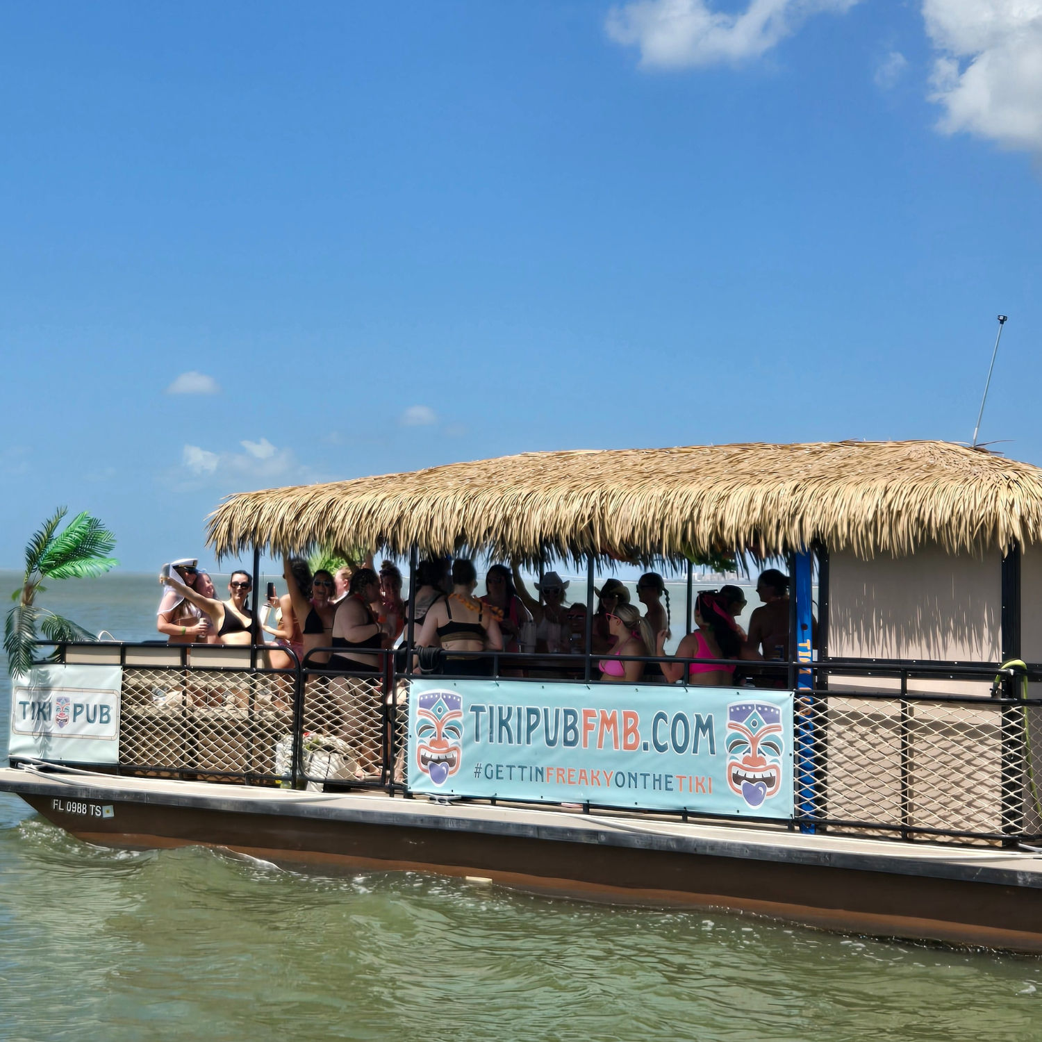 Tiki-style party boat with thatched roof and people in swimsuits cruising calm coastal Florida waters under a bright blue sky