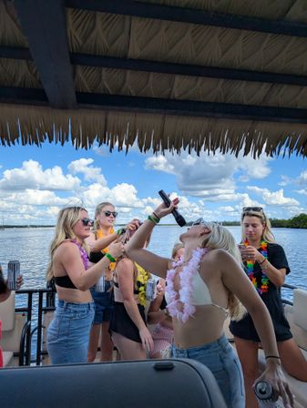 Group of women partying on a tiki-roofed pontoon boat on a lake, wearing leis and swimsuits, singing into a microphone and holding drinks under a bright blue sky with puffy clouds.