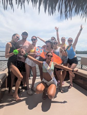 Group of friends celebrating a sunny tropical boat party on the water, wearing swimsuits and a bachelorette sash while holding a colorful tiki sign under a thatched canopy