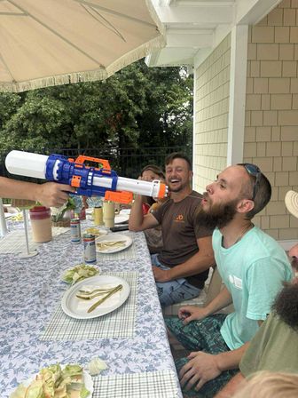 Friends at a backyard patio table laughing as someone sprays a large blue-and-orange water blaster into a bearded man's mouth under a patio umbrella during a summer outdoor meal.