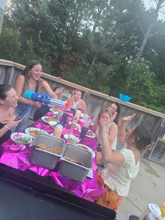 Friends at a backyard patio dinner, laughing around a pink-covered picnic table with food trays and condiments as one playfully aims a blue water gun.