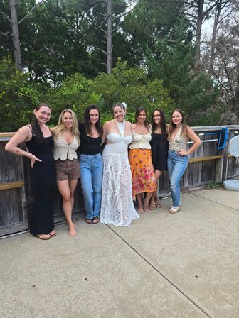 Seven friends smiling on a wooden deck and concrete patio with pine trees behind, center woman in a white lace dress and others in casual summer outfits.