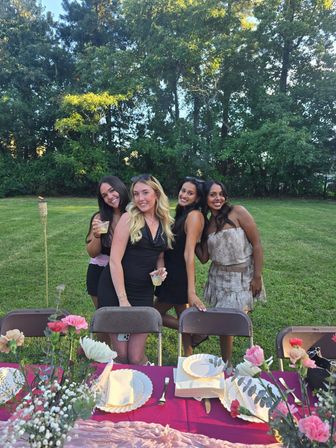 Four smiling friends posing behind chairs at a backyard summer dinner party with a pink floral tablescape, drinks, tiki torch, and tree-lined lawn