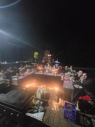 Nighttime backyard patio party on a paved patio — guests around a U-shaped table lit by string lights and candles, raising glasses; grill and condiment bottles in the foreground, patio heater and gold '2' balloon in the background.