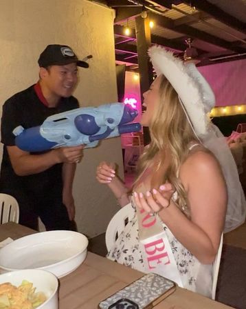 Bride-to-be wearing a white veil and sash laughing as a person playfully aims a large blue water gun at her on a lit outdoor patio during a bachelorette party