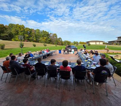 Fun suburban backyard dinner on a spacious patio: U-shaped long tables with family and friends seated near a pool and wide lawn, autumn trees and a blue sky with scattered clouds.