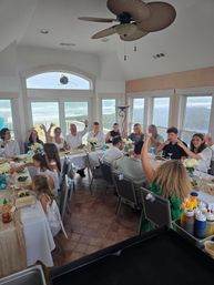 Cheerful beachfront family gathering in a bright ocean-view dining room — long table with white floral centerpieces and guests raising a toast.