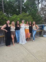 Seven friends smiling on a wooden deck and concrete patio with pine trees behind, center woman in a white lace dress and others in casual summer outfits.