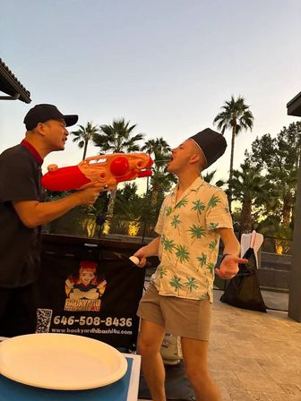 Playful outdoor patio scene at sunset with palm trees: a person in a black cap aims a large red water gun at another wearing a chef hat and tropical palm-print shirt who playfully opens their mouth.