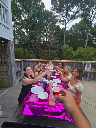 Group of friends toasting with small red cups around a shiny pink-covered table on a wooden outdoor deck surrounded by tall pine trees.
