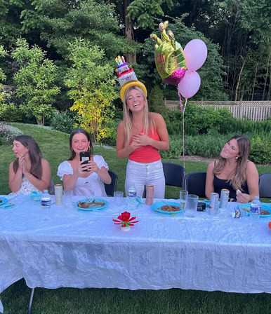 Smiling group of friends at a backyard garden birthday celebration: woman standing in a novelty cake hat with balloons behind her, three friends seated at a long white‑clothed table with plates and drinks.