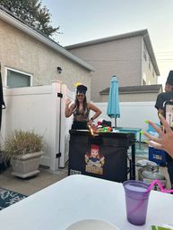 Backyard patio party scene with a woman in a novelty hat and sunglasses waving beside a grill with a small flame, white vinyl fence, turquoise umbrella, propane tank and a purple cup in the foreground.