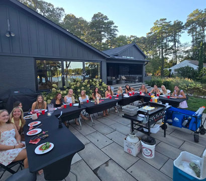 Group of friends smiling around U-shaped black-covered tables on a stone backyard patio for a summer lakeside dinner party, red cups and plates, portable grill and coolers nearby, modern dark-gray house and tall pine trees in the background.