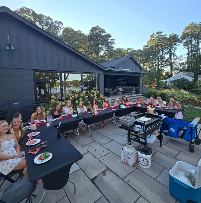 Group of friends smiling around U-shaped black-covered tables on a stone backyard patio for a summer lakeside dinner party, red cups and plates, portable grill and coolers nearby, modern dark-gray house and tall pine trees in the background.