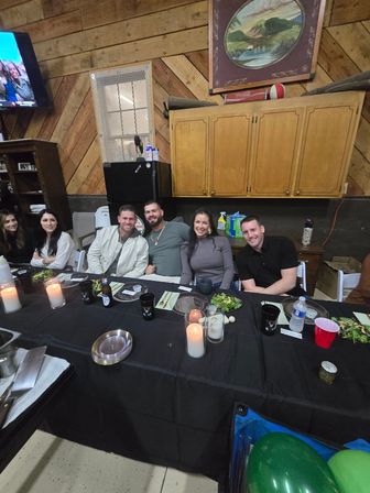 Group of friends smiling at a long black-clothed table in a rustic wood-paneled event space, with candles, salads, drinks and green balloons for a casual dinner celebration.